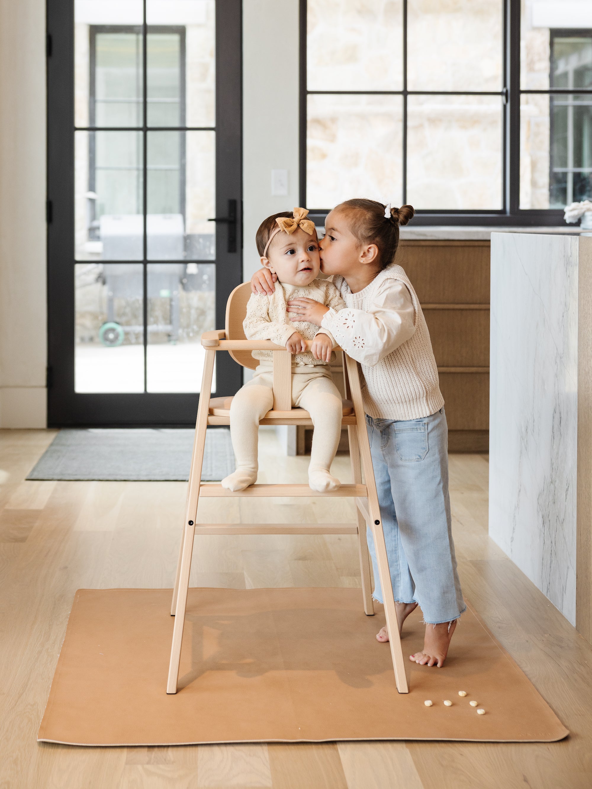 Baby in highchair with sibling kissing them on the cheek.
